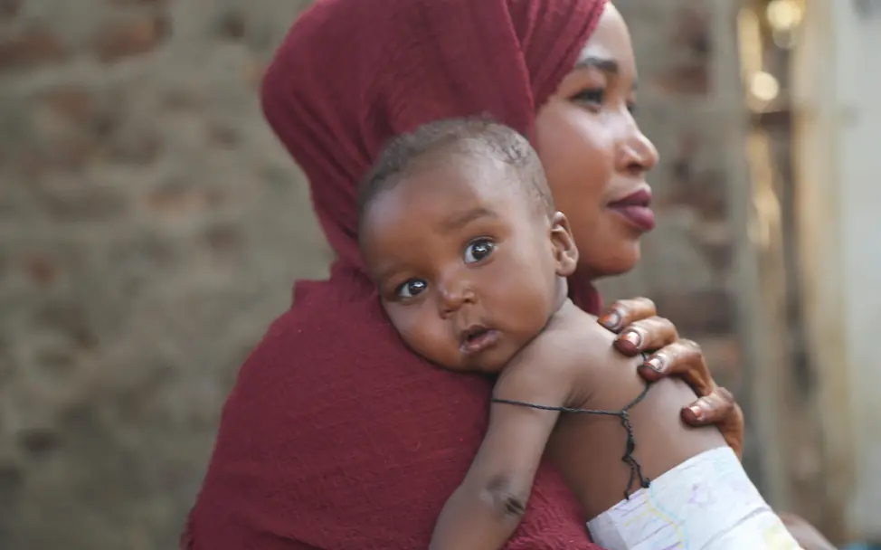 Mangelernährung im Sudan | © UNICEF/UNI593639/ Eine Mutter hat ihr Kleinkind auf dem Arm welches in die Kamera schaut